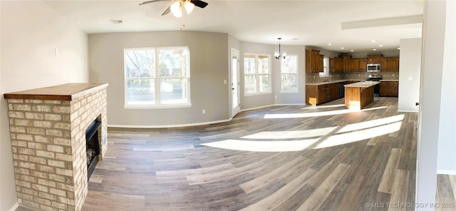 unfurnished living room featuring visible vents, a brick fireplace, baseboards, ceiling fan with notable chandelier, and wood finished floors