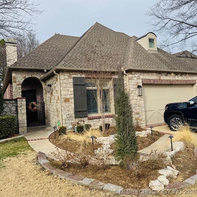 french country home featuring an attached garage, stone siding, and roof with shingles