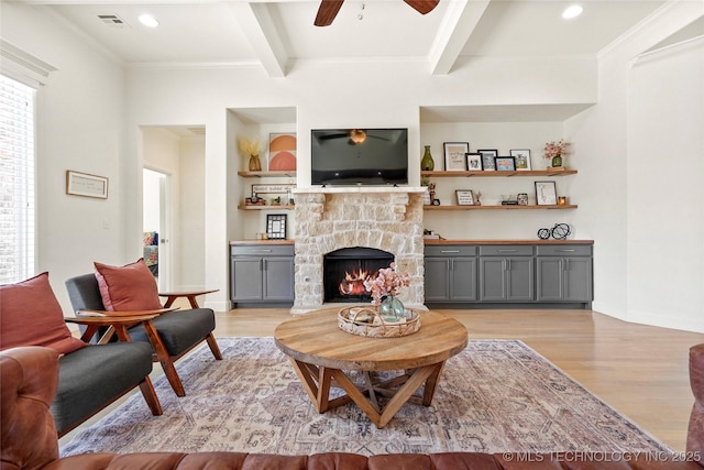 living room featuring light wood finished floors, crown molding, beamed ceiling, a stone fireplace, and a ceiling fan