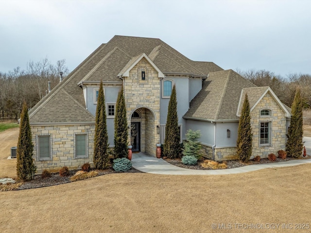 french country inspired facade with stucco siding, central air condition unit, and a shingled roof