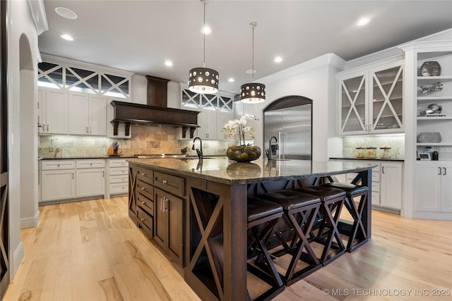 kitchen with built in refrigerator, light wood-type flooring, custom range hood, and a kitchen island with sink