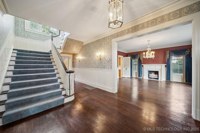 unfurnished living room with wood finished floors, a wainscoted wall, wallpapered walls, a warm lit fireplace, and a notable chandelier