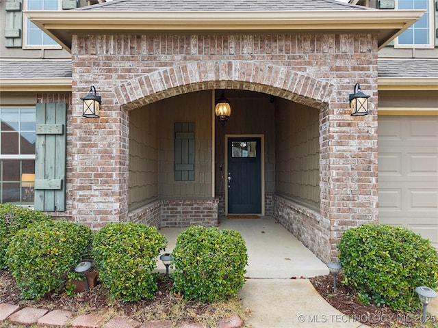 doorway to property featuring an attached garage, brick siding, and roof with shingles