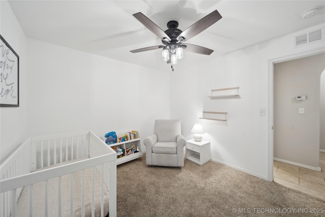 carpeted bedroom with a crib, baseboards, visible vents, and ceiling fan