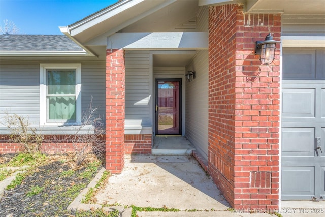 entrance to property featuring a garage, brick siding, and roof with shingles