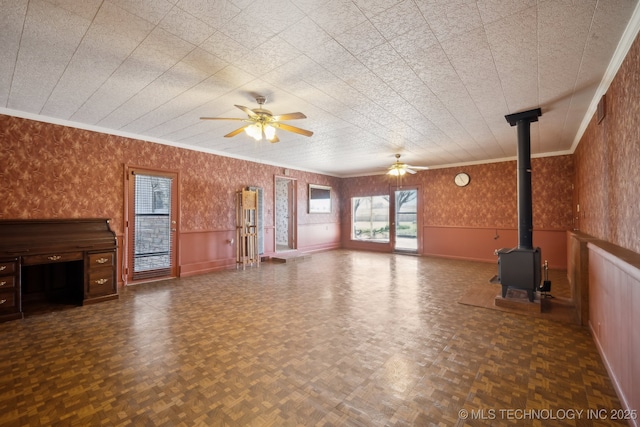 unfurnished living room featuring a wainscoted wall, a ceiling fan, crown molding, wallpapered walls, and a wood stove