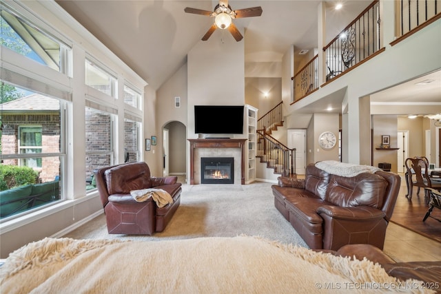 living area featuring high vaulted ceiling, a ceiling fan, a glass covered fireplace, stairway, and baseboards