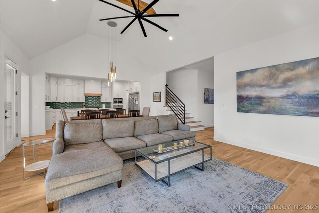 living area featuring baseboards, ceiling fan, stairs, light wood-style floors, and high vaulted ceiling