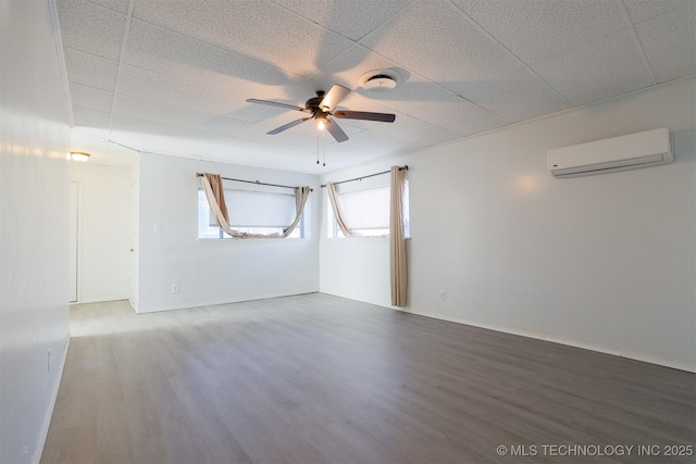 spare room with baseboards, a drop ceiling, a wall mounted air conditioner, wood finished floors, and a ceiling fan