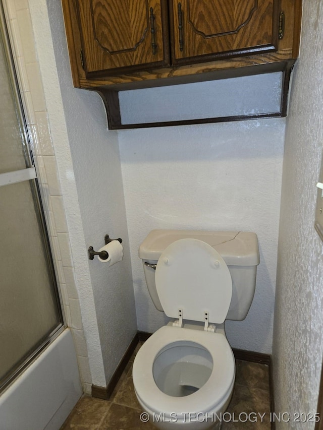 bathroom featuring baseboards, tile patterned flooring, toilet, a textured wall, and combined bath / shower with glass door