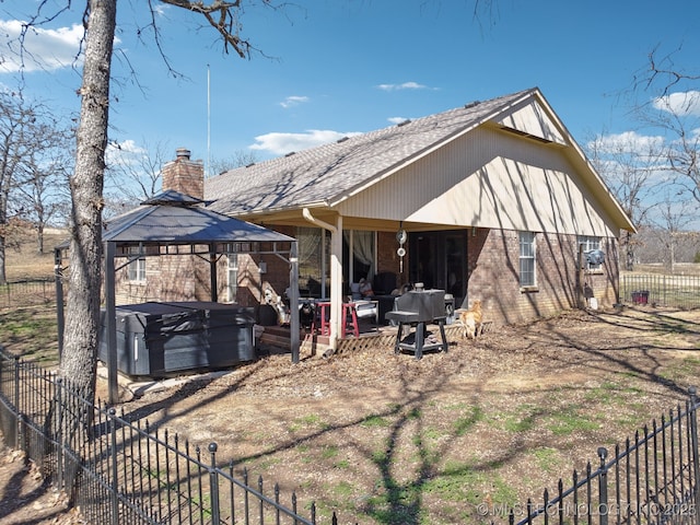 rear view of house with brick siding, a hot tub, fence, a gazebo, and a chimney