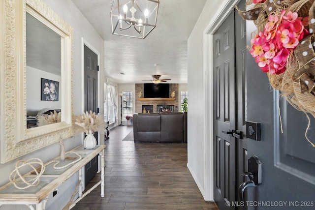 foyer entrance featuring baseboards, dark wood finished floors, a textured ceiling, a brick fireplace, and ceiling fan with notable chandelier