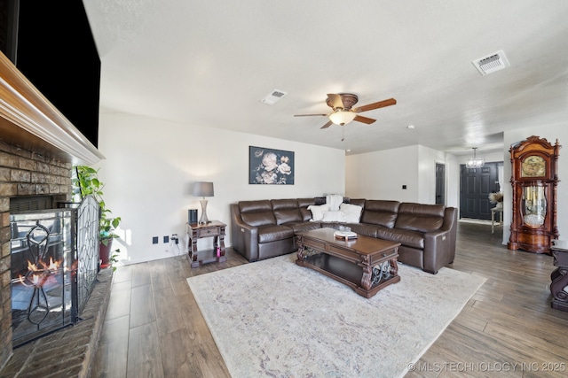 living room with visible vents, a brick fireplace, wood finished floors, and ceiling fan with notable chandelier