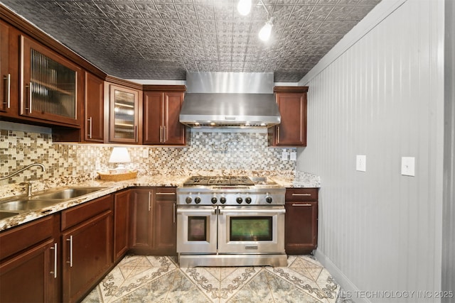 kitchen featuring a sink, double oven range, tasteful backsplash, wall chimney range hood, and glass insert cabinets