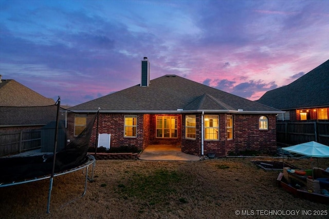 back of house at dusk with a trampoline, fence, brick siding, a chimney, and a patio area