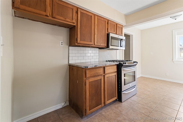 kitchen featuring dark stone countertops, backsplash, appliances with stainless steel finishes, light tile patterned flooring, and brown cabinetry