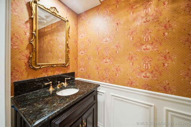 bathroom with vanity, a decorative wall, wainscoting, and wallpapered walls