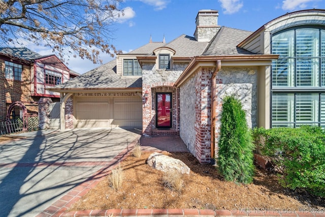 view of front of property featuring driveway, stone siding, an attached garage, a shingled roof, and a chimney