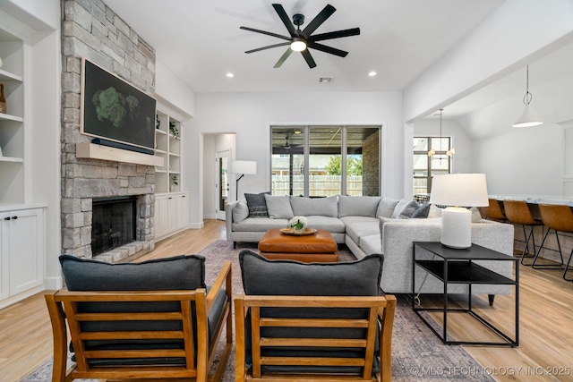 living area featuring a stone fireplace, light wood-style flooring, ceiling fan with notable chandelier, and built in features