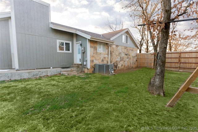 rear view of house with entry steps, fence private yard, central AC unit, a yard, and stone siding