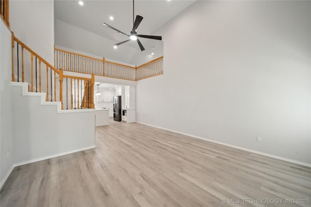 unfurnished living room with a ceiling fan, baseboards, stairs, a towering ceiling, and light wood-type flooring