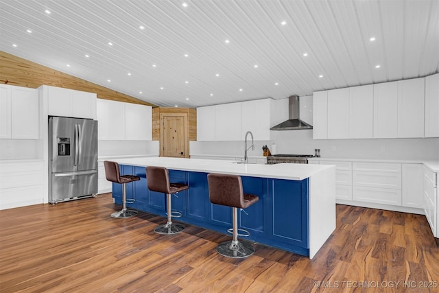 kitchen with a spacious island, wall chimney range hood, dark wood finished floors, stainless steel fridge, and a sink