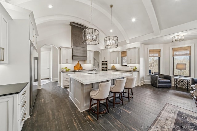 kitchen featuring custom exhaust hood, an island with sink, arched walkways, stainless steel appliances, and a sink