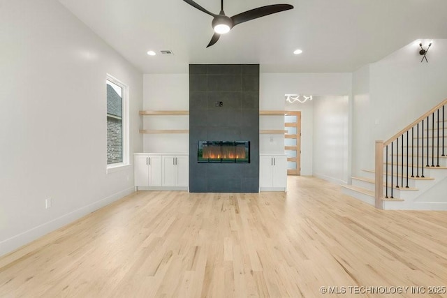 unfurnished living room with a tiled fireplace, stairway, light wood-type flooring, and ceiling fan