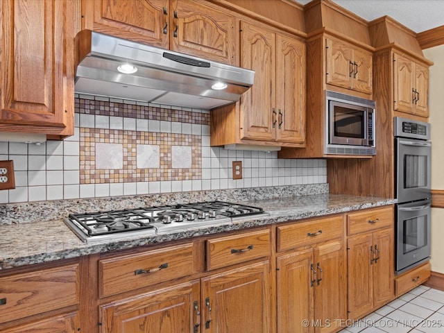 kitchen with under cabinet range hood, appliances with stainless steel finishes, tasteful backsplash, and light stone countertops