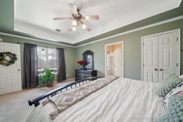 carpeted bedroom with a ceiling fan, visible vents, a tray ceiling, ensuite bathroom, and crown molding