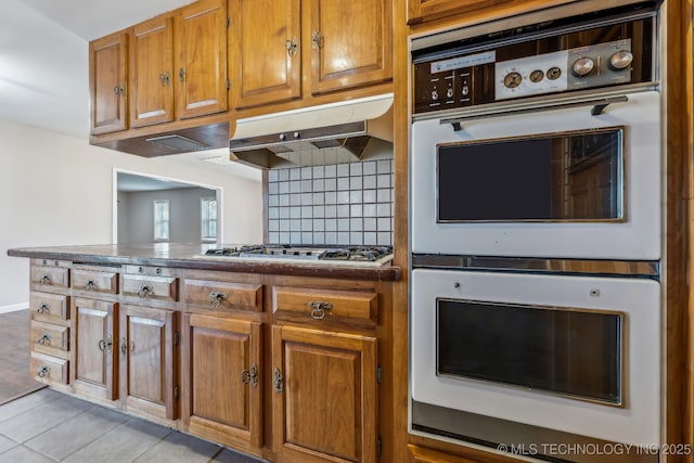 kitchen with gas cooktop, brown cabinetry, white double oven, under cabinet range hood, and tasteful backsplash