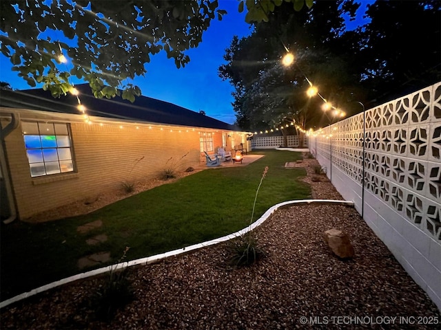 property exterior at night featuring brick siding, a patio area, a lawn, and a fenced backyard