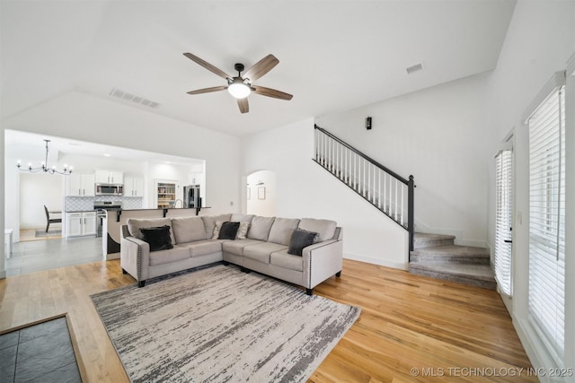 living area with stairway, a ceiling fan, light wood finished floors, a chandelier, and vaulted ceiling