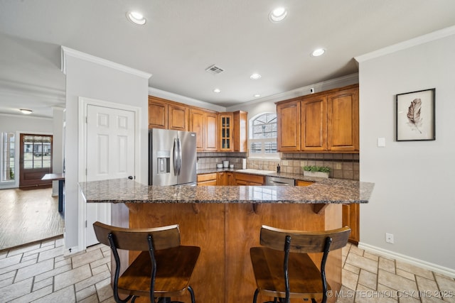 kitchen with backsplash, stone tile floors, appliances with stainless steel finishes, brown cabinetry, and crown molding