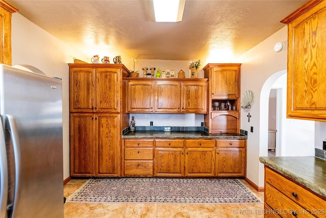 kitchen featuring dark countertops, arched walkways, freestanding refrigerator, and brown cabinets