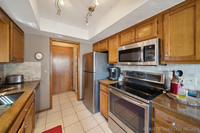 kitchen featuring a tray ceiling, decorative backsplash, brown cabinets, appliances with stainless steel finishes, and light tile patterned flooring