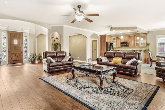 living room featuring baseboards, light wood-type flooring, ornamental molding, arched walkways, and a ceiling fan