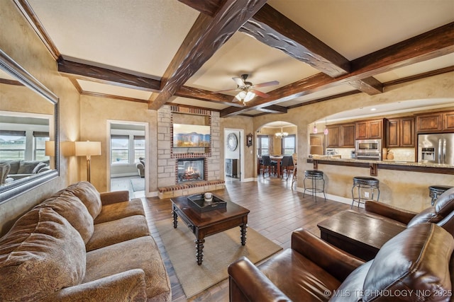 living room featuring wood finished floors, baseboards, arched walkways, a brick fireplace, and beamed ceiling