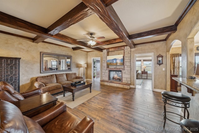 living room featuring a brick fireplace, baseboards, beam ceiling, arched walkways, and wood-type flooring