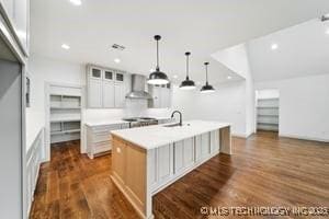 kitchen with a center island with sink, light countertops, dark wood-style flooring, wall chimney exhaust hood, and a sink