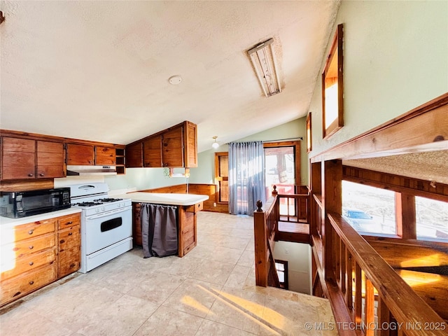 kitchen featuring black microwave, gas range gas stove, under cabinet range hood, vaulted ceiling, and brown cabinetry