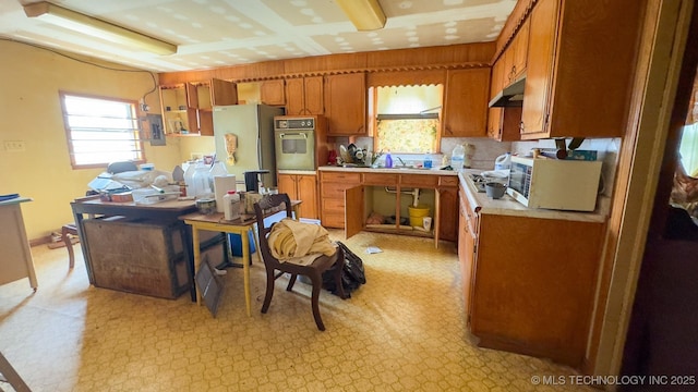 kitchen featuring under cabinet range hood, light countertops, brown cabinets, freestanding refrigerator, and white oven