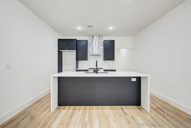 kitchen featuring light wood finished floors, wall chimney range hood, an island with sink, light countertops, and decorative backsplash