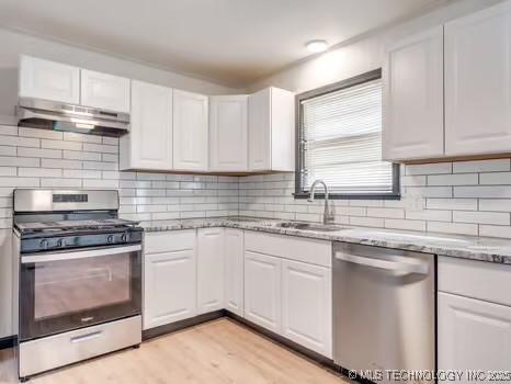 kitchen featuring under cabinet range hood, light stone counters, a sink, appliances with stainless steel finishes, and white cabinets