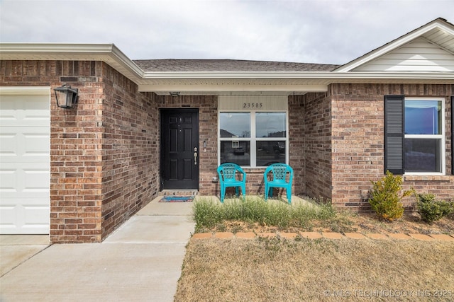 view of exterior entry with brick siding, a garage, and roof with shingles