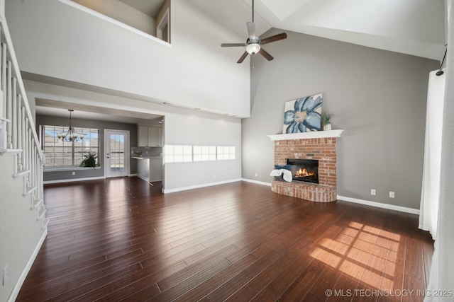 unfurnished living room with ceiling fan with notable chandelier, dark wood-style floors, baseboards, a brick fireplace, and stairs