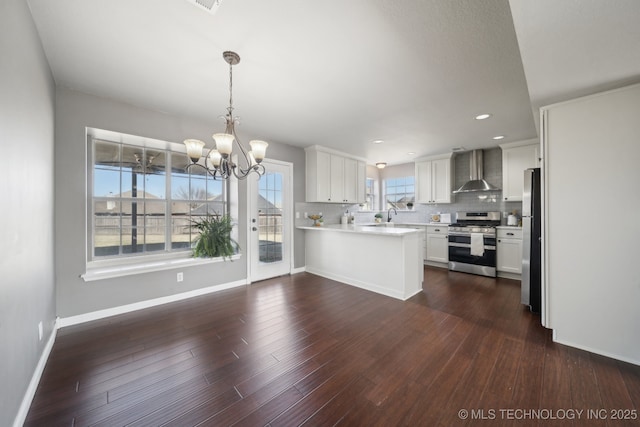 kitchen featuring a peninsula, decorative backsplash, appliances with stainless steel finishes, wall chimney exhaust hood, and a chandelier