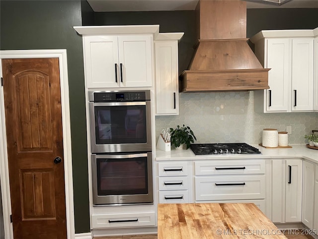 kitchen with decorative backsplash, appliances with stainless steel finishes, wood counters, custom range hood, and white cabinetry