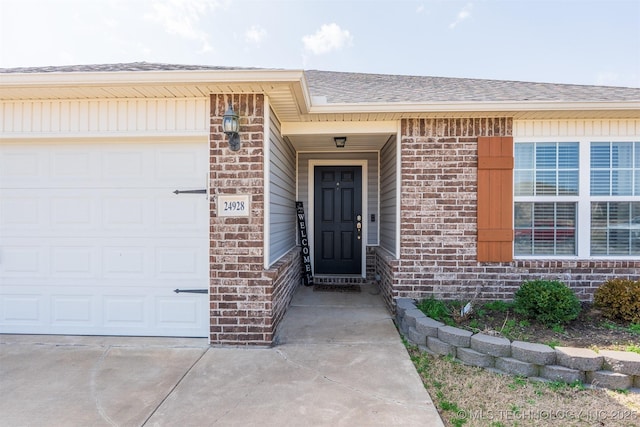 view of exterior entry with a garage, brick siding, driveway, and a shingled roof