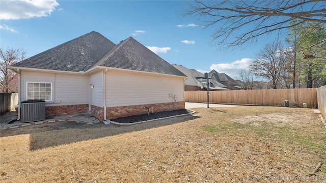 view of side of property with roof with shingles, central AC unit, a fenced backyard, and a lawn